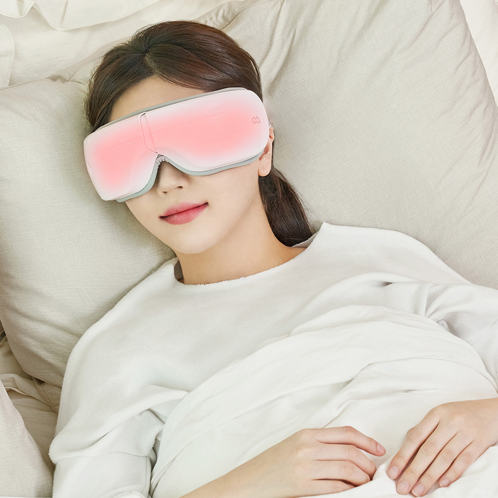 분홍색 온열 패턴이 보이는 안마기를 쓴 여성이 침대에 누워 편안한 표정으로 있는 장면