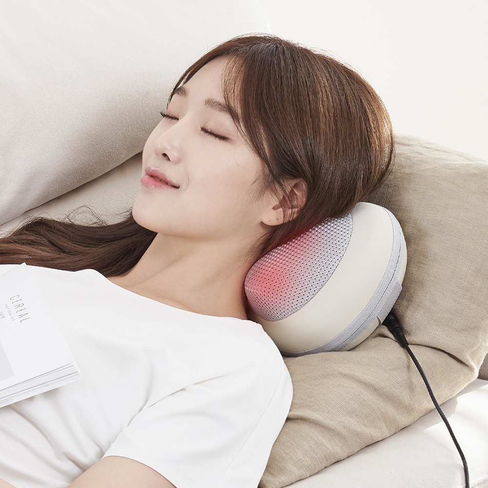 여성이 소파에 누워 목과 어깨에 핑크색 라인이 있는 크림색 마사지기를 착용하고 편안한 표정으로 휴식하는 장면