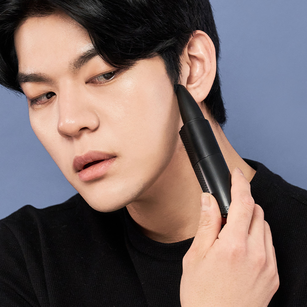 남성이 검은 셔츠를 입고 파란 배경 앞에서 검은색 펜슬 형태의 트리머를 귀 옆에 들고 있는 모습