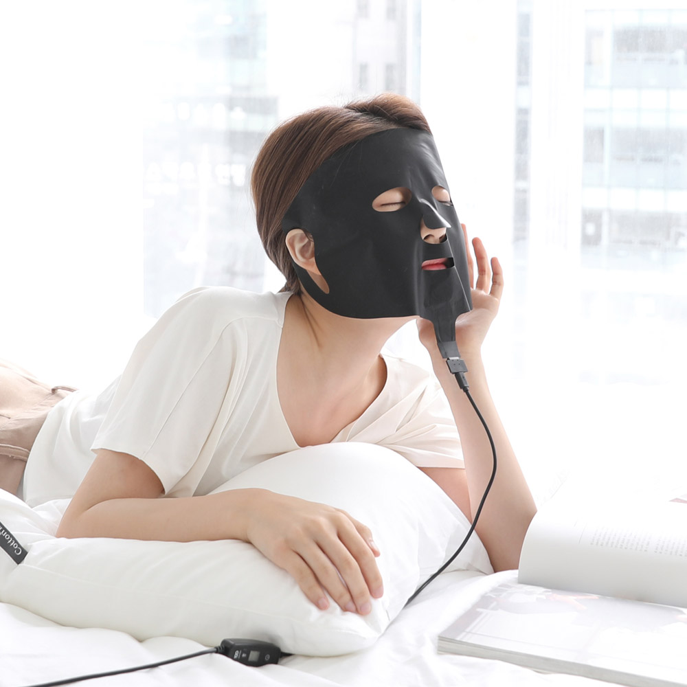 침대에 누워 검은색 마스크를 착용하고 안경알 같은 패드 부분을 손으로 고정하며 온열 기능을 사용 중인 모습