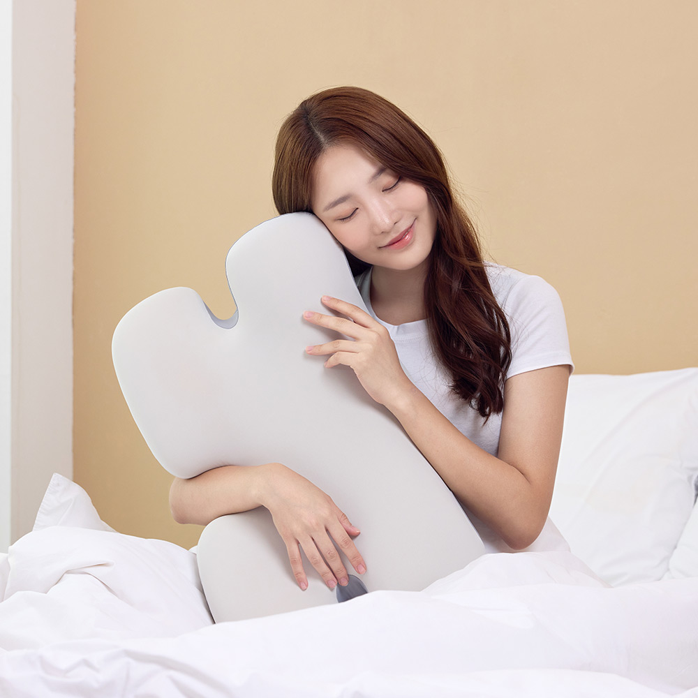 침대에 앉아 흰색 경추 베개 커버를 손으로 만지며 편안함을 느끼는 여성