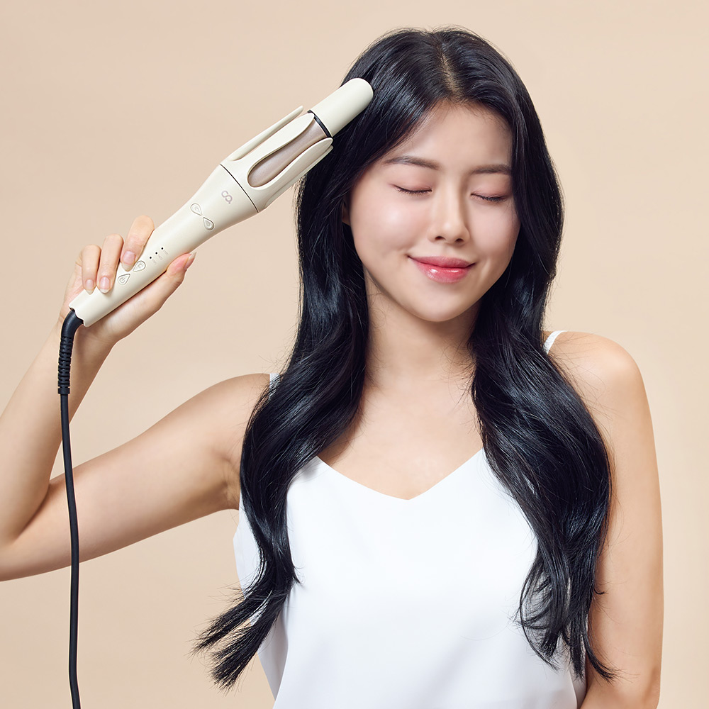 베이지색 자동 회전 고데기를 옆머리에 대고 있는 여성의 얼굴과 긴 머리