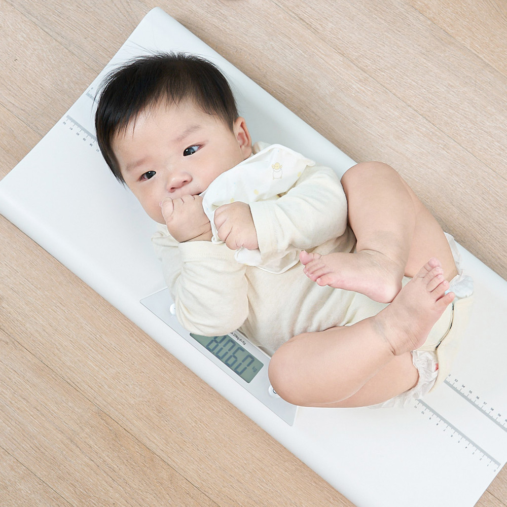 하얀 체중계 위에 누운 신생아의 몸무게와 키를 측정하는 장면