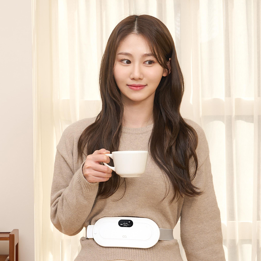 서 있는 자세에서 여성의 복부에 부착된 흰색 온열 찜질기와 디지털 디스플레이가 보이는 모습