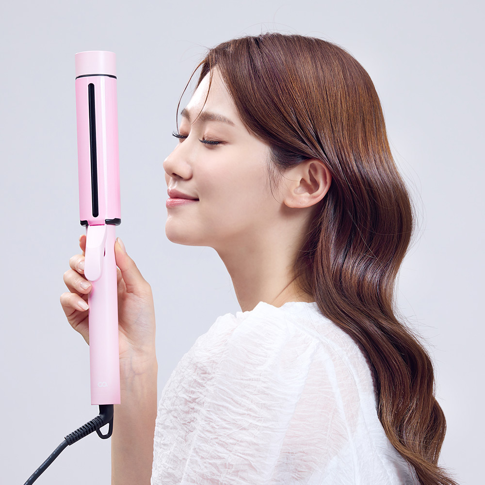 핑크색 고데기를 옆으로 들어 보이는 여성이 완성된 웨이브 머리의 옆 프로필을 보여주는 장면