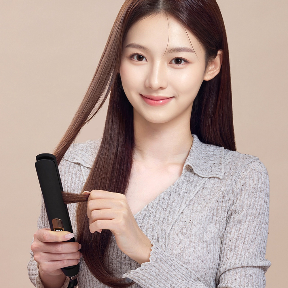 긴 갈색 머리를 가진 여성이 회색 옷을 입고 고데기로 머리를 스타일링하는 모습