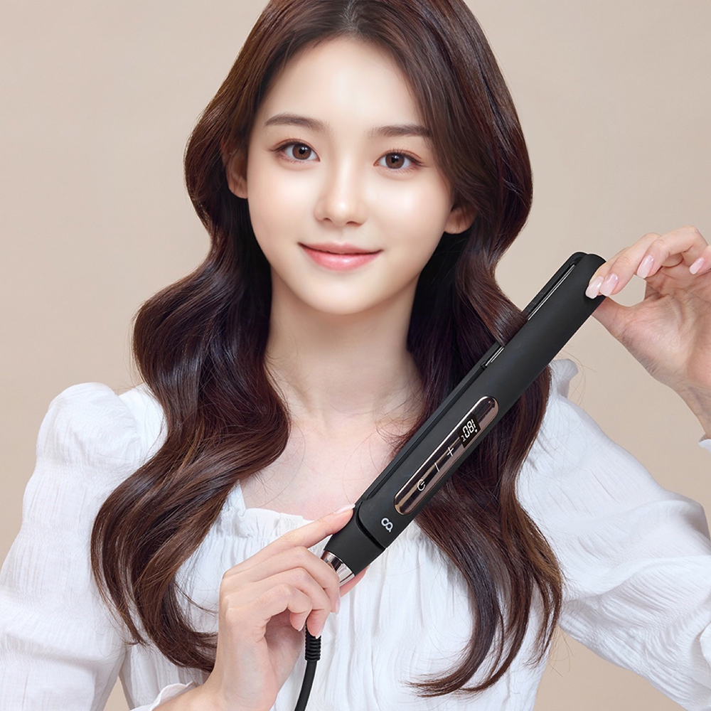 흰색 옷을 입은 여성이 손에 고데기를 들고 웨이브 펌 스타일이 완성된 갈색 머리를 보여주는 모습