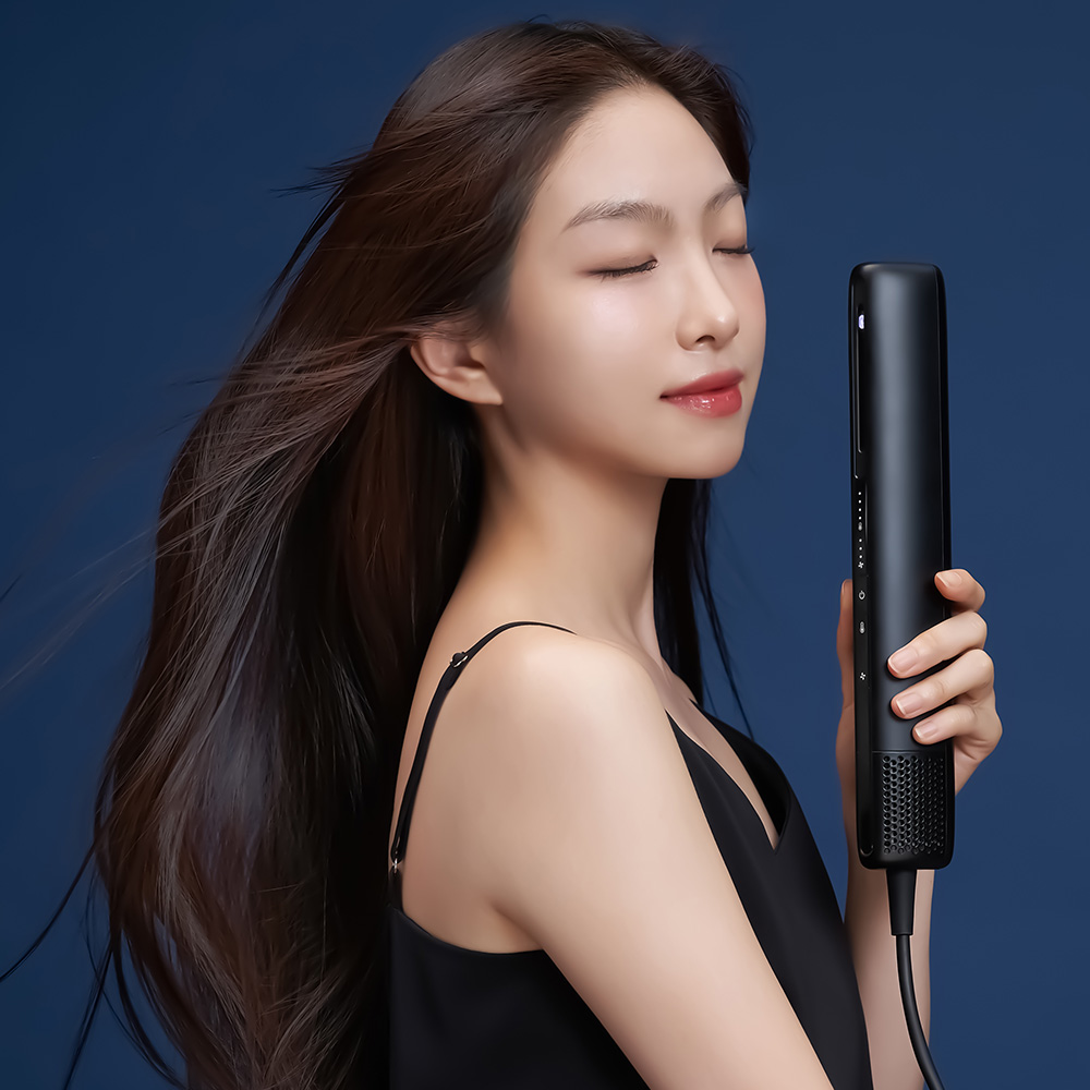 검은색 슬림 드라이기를 손에 들고 있는 여성이 긴 검은 머리를 옆으로 흘러내린 모습
