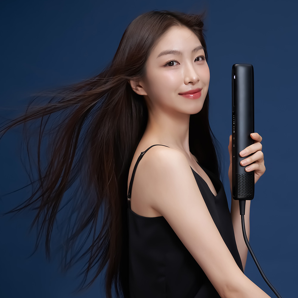 검은색 원형 드라이기를 들고 있는 여성의 옆모습으로 공기 흐름에 머리가 날리는 모습