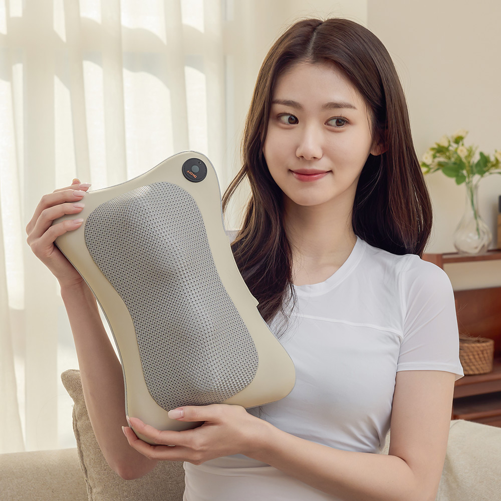 앉은 자세에서 마사지 쿠션을 옆으로 들어 보이며 미소 짓는 여성의 모습