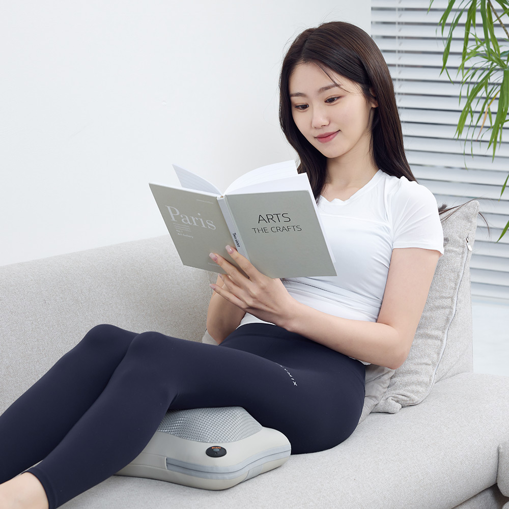 소파에 기대어 책을 읽으면서 마사지 쿠션을 허리 아래에 두고 사용하는 모습