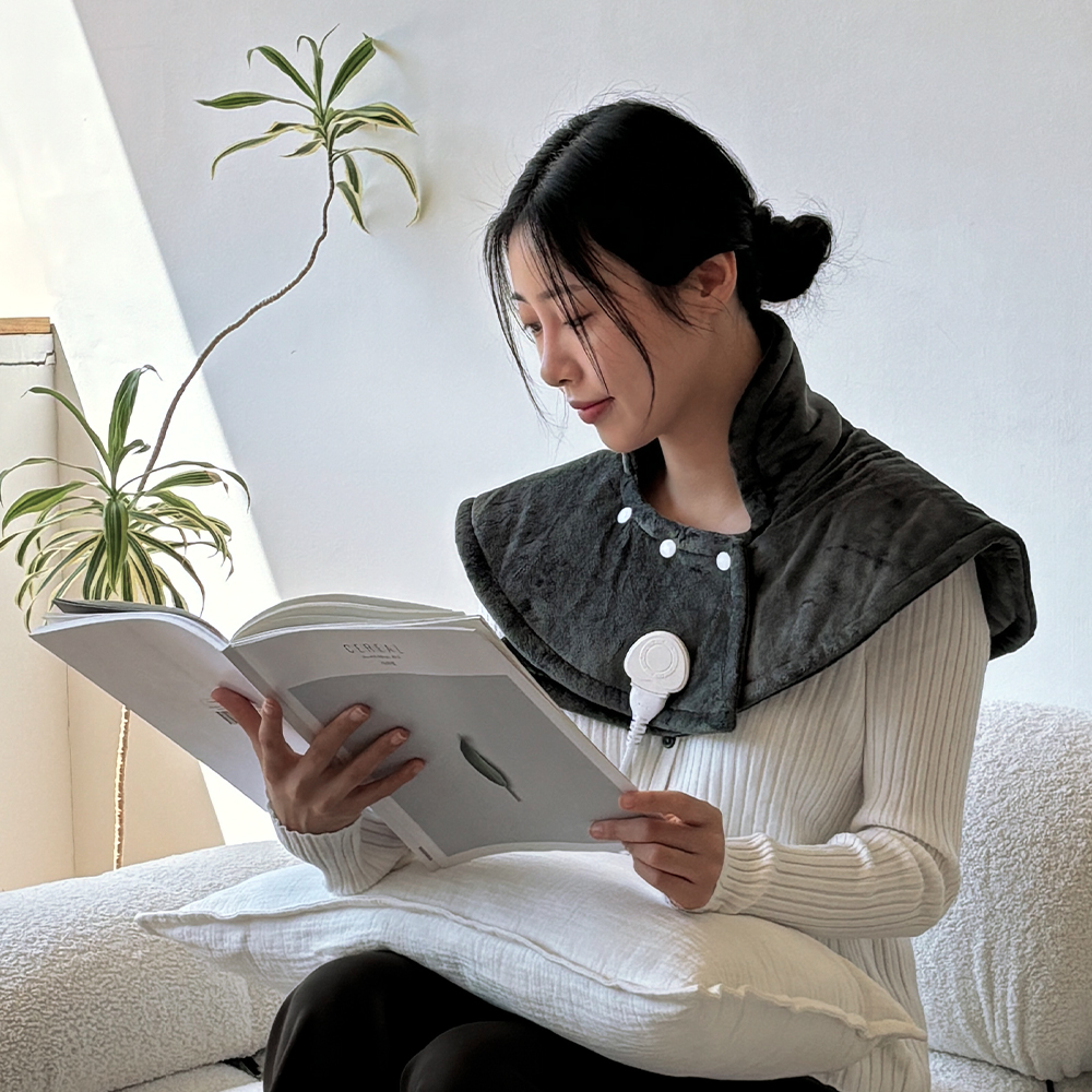 밝은 실내에서 책을 읽으며 검은색 어깨 찜질기를 착용한 여성의 정면 모습