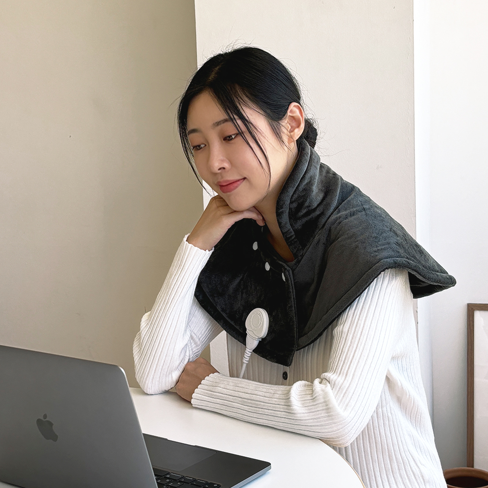 검은색과 회색 투톤의 어깨 찜질기를 착용한 여성이 데스크에서 노트북 앞에 앉아 있는 모습