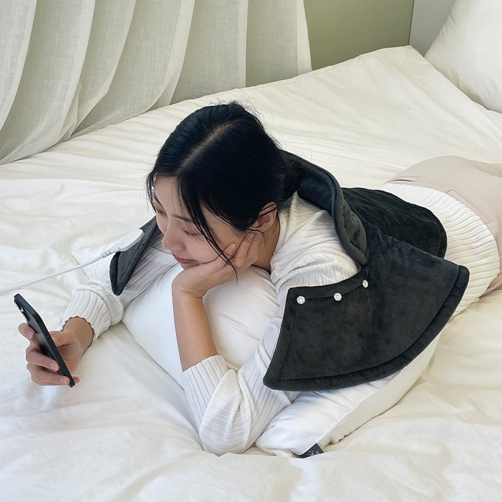 침대에서 온열 찜질기를 어깨에 걸친 채 스마트폰을 들고 있는 사람의 모습
