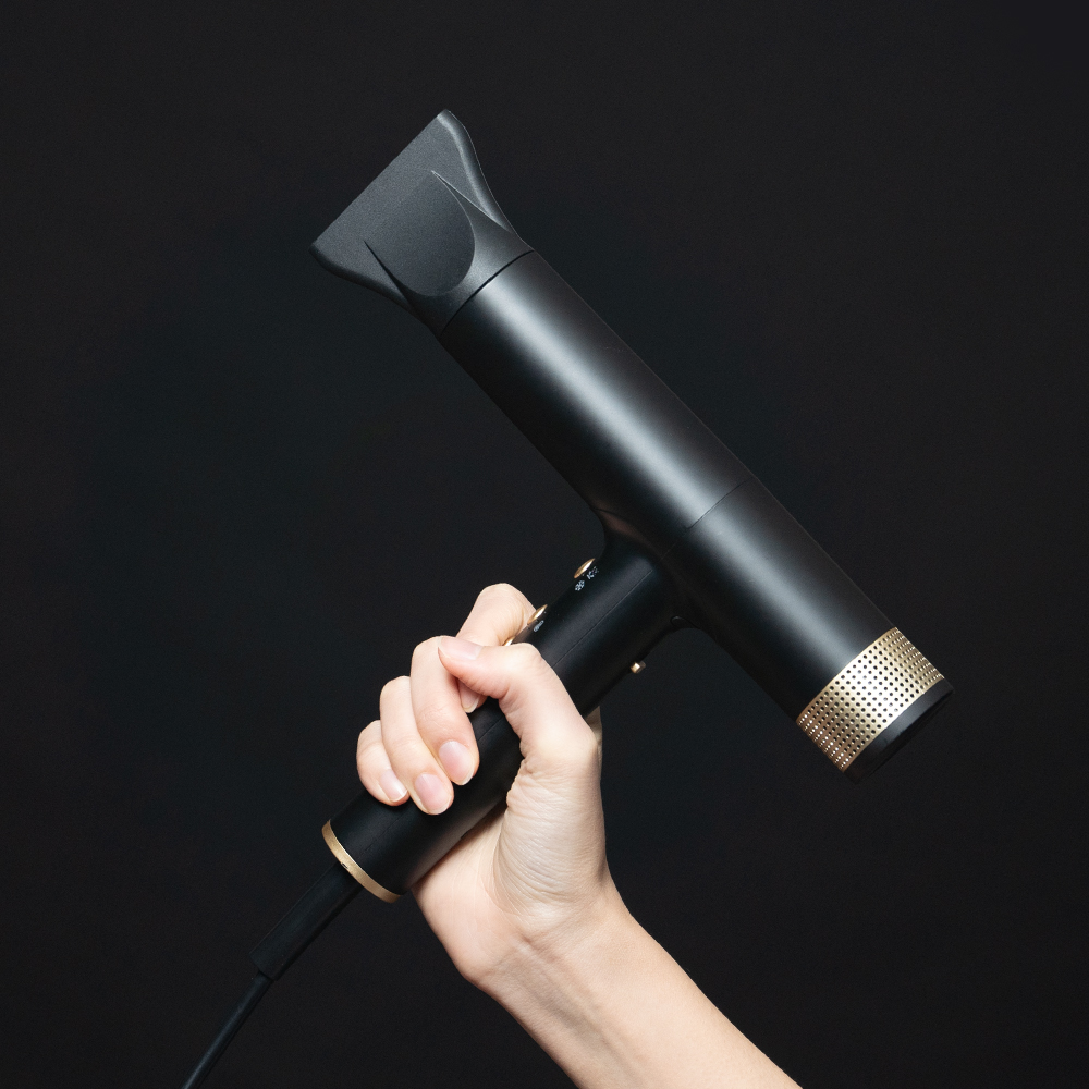 검은색과 금색으로 디자인된 슬림한 드라이기를 손에 들고 위쪽을 향해 들어 올린 모습