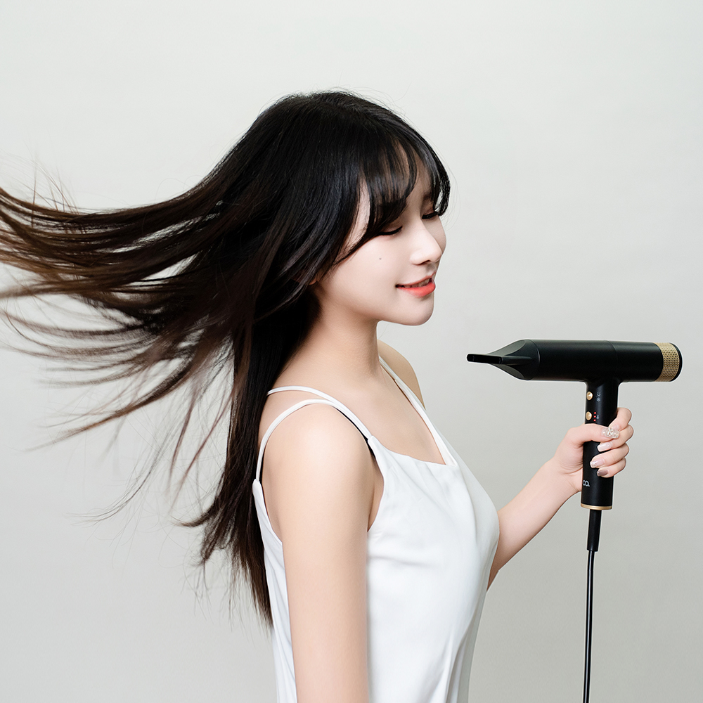 여성이 하얀 민소매 상의를 입고 검은색 드라이기를 사용하며 긴 머리가 바람에 움직이는 사용 장면을 측면에서 촬영한 이미지