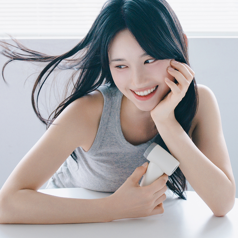 흰색 미니 선풍기를 손에 들고 있는 여성이 웃으면서 얼굴 옆에서 바람을 쐬고 있는 장면