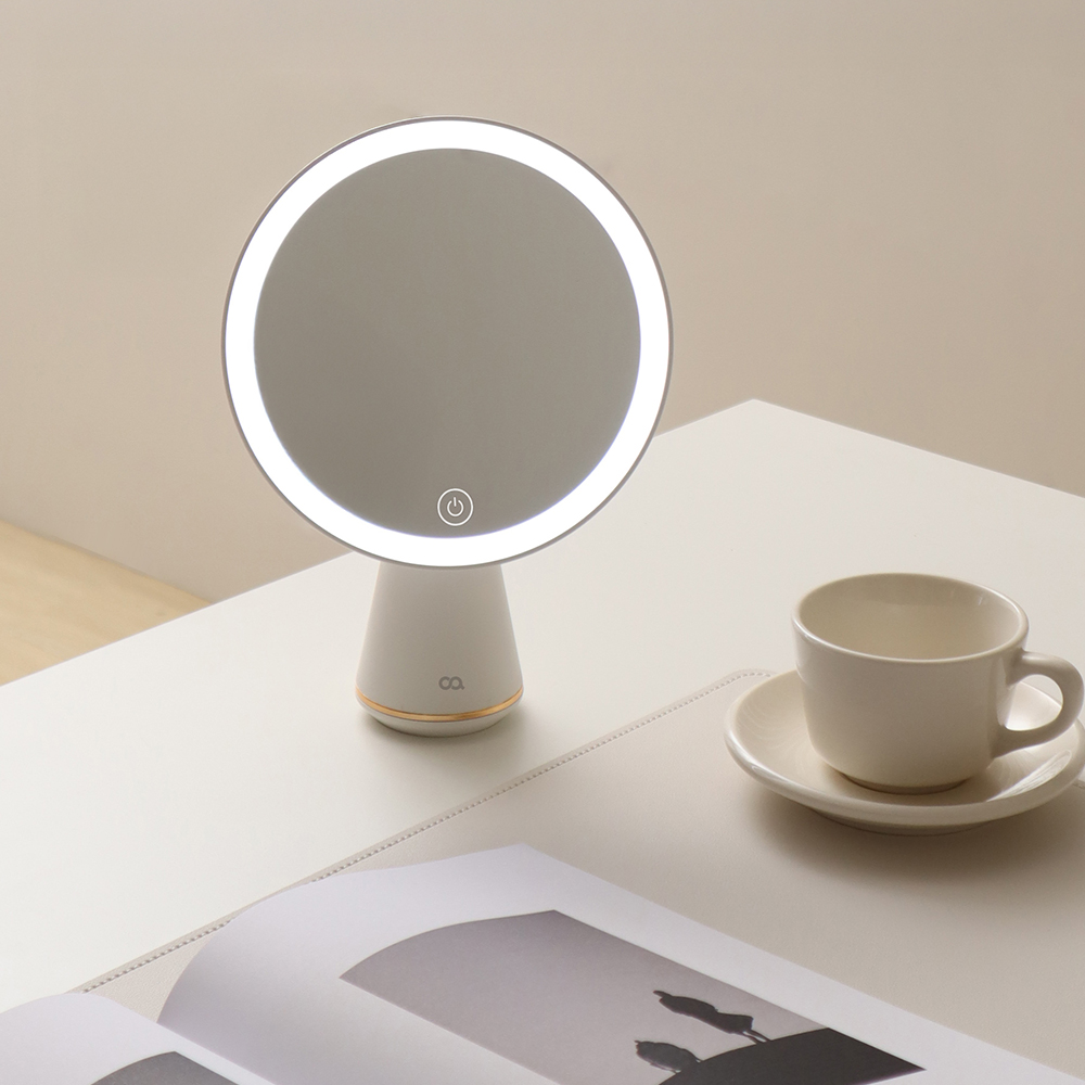 베이지톤 테이블 위에 세워진 원형 LED 거울 옆에 흰색 커피잔과 잡지가 놓여 있는 일상적인 공간 구성