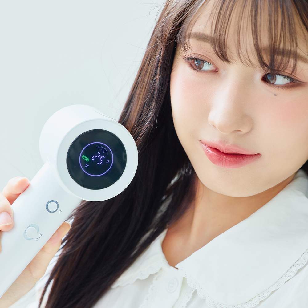 여성이 흰색 헤어 드라이기를 손에 들고 얼굴 옆에 보이며 제품의 원형 LED 디스플레이와 컨트롤 버튼을 정면으로 보여주는 사진