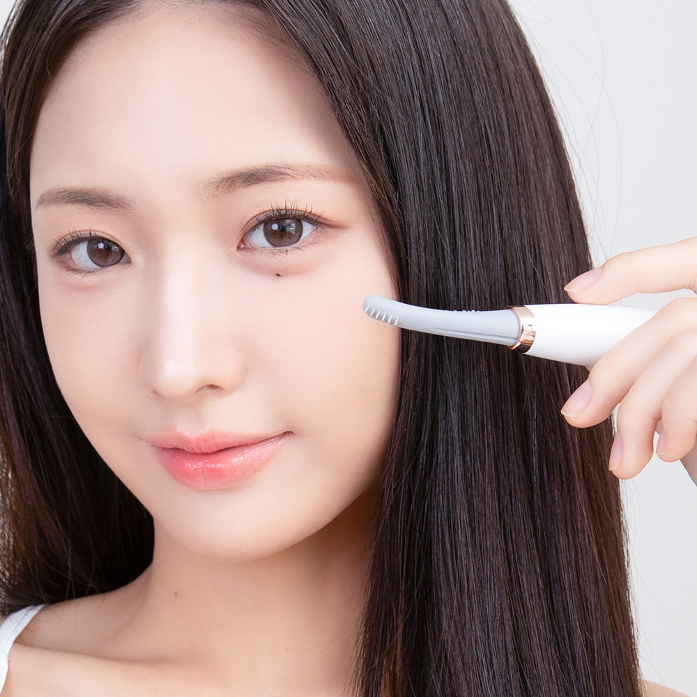 여성이 흰색 속눈썹 고데기로 눈썹을 올리는 모습을 정면에서 촬영한 이미지