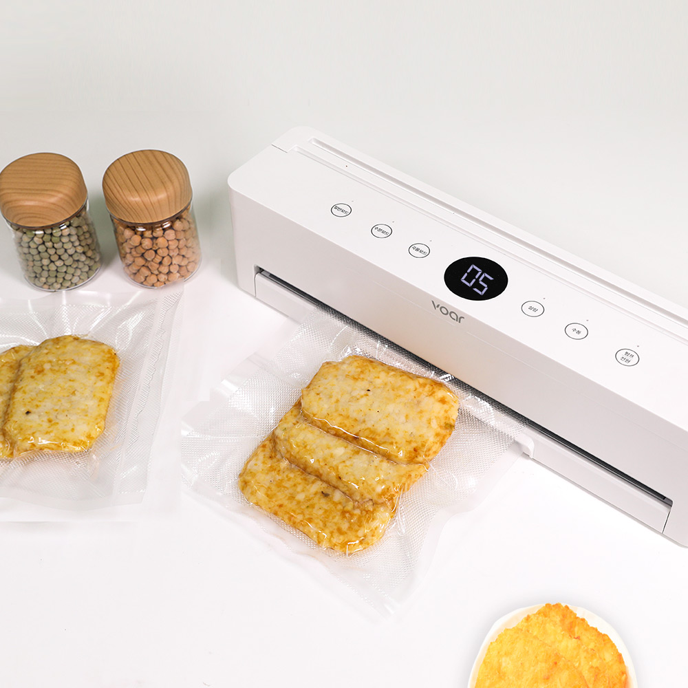 흰색 진공포장기 앞에 밥과 반찬이 투명한 진공 비닐에 포장되어 있으며, 곡물 통이 배경에 놓인 상태