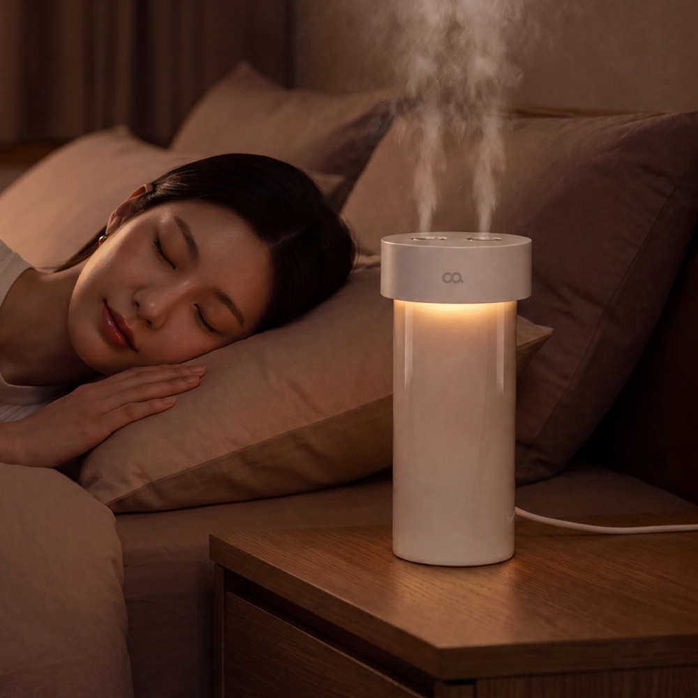 침대에 누워 있는 여성의 옆 테이블 위에 베이지색 가습기가 놓여 있고, 따뜻한 무드등 조명과 함께 수증기가 피어올라 야간 실내 환경을 촉촉하게 유지하는 모습이다.