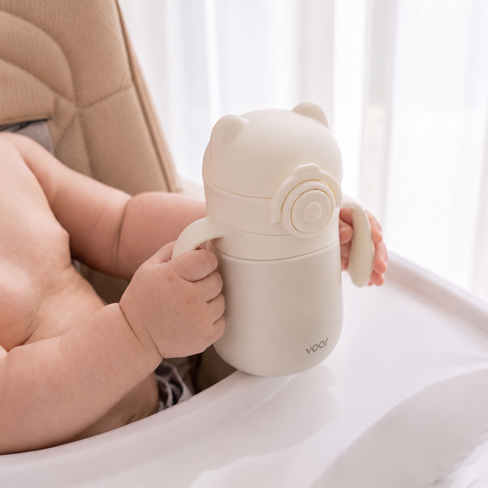 창가의 밝은 자연광 아래서 어른 손에 쥐어진 곰 모양 빨대컵의 옆모습으로 아기 손 크기에 맞는 컵의 사이즈를 보여주는 사진
